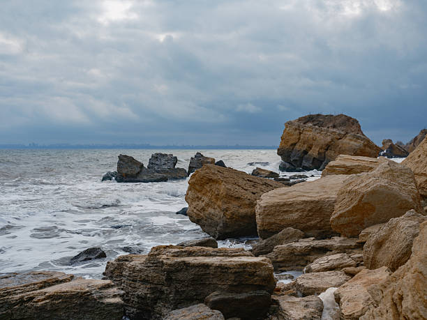 Wild Black Sea coast landscape with layers of eroded boulders and rocks on the shore. Powerful waves, sea foam, moody dark sky, distant city on horizon, coastal erosion, autumn.