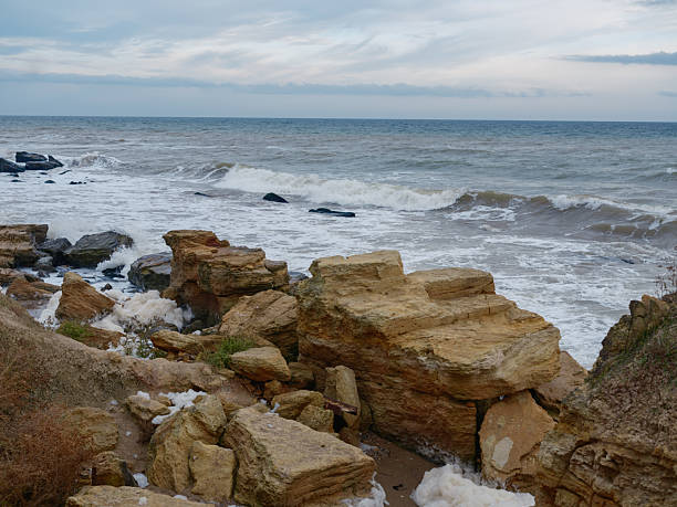 Wild Black Sea coast with prominent layered sandstone rocks in foreground. Strong waves, sea foam, moody horizon, cloudy sky, geological feature, cold nature landscape.