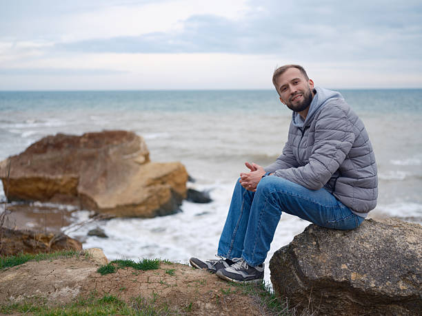 Smiling attractive man sitting on a rock by the wild Black Sea coast. Casual grey jacket, jeans, peaceful travel, beautiful nature background, autumn vacation.