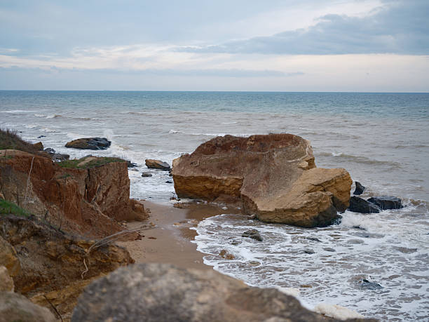 Wild Black Sea coast landscape with large yellow-brown rock and steep clay cliff. Waves breaking on shore, moody sky, coastal erosion, natural beauty, travel destination.