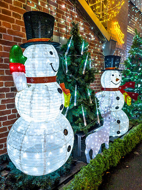 Two illuminated snowmen wearing top hats and holding gift boxes stand before a lit brick facade; a lighted reindeer and decorated fir trees create a warm, festive atmosphere