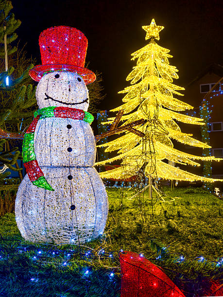 istock A large illuminated snowman figure stands before a golden lit, stylized Christmas tree on a market square. String lights, garlands and a wire reindeer complete the festive nighttime decoration. 2250290945