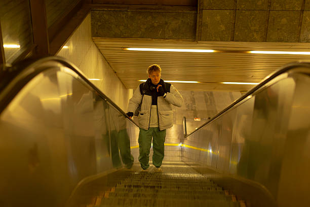 Caucasian man on escalator in subway descending alone toward concourse, wearing puffer jacket and patterned backpack, warm yellow lighting, reflective metal handrails, candid documentary mood,