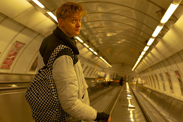 Caucasian man on walkway in metro turning toward camera with intent gaze, patterned backpack slung over shoulder, long tunnel of fluorescent lights, reflective floor, candid urban portrait, steady