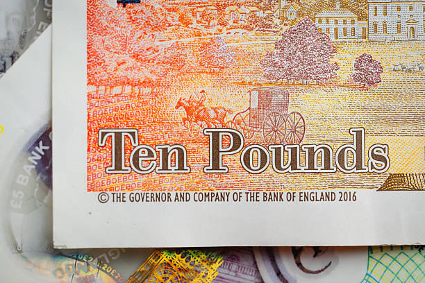 A close-up view of a ten-pound banknote highlighting its detailed printed illustration, including warm orange and yellow tones, fine linework, and classic carriage imagery. The composition emphasizes texture, color gradients, and traditional currency design elements. No property release is available for the depiction of official banknotes.