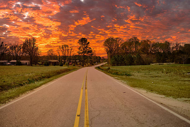 Outdoor scene of a rural road in the southern United States during sunset with orange and red sky, surrounded by green fields and sparse trees, creating a peaceful, natural setting in early evening