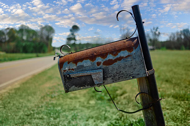 A rusty metal mailbox mounted on a black post beside a narrow rural road with grassy fields and blurred tree line under a partly cloudy sky in the southern countryside