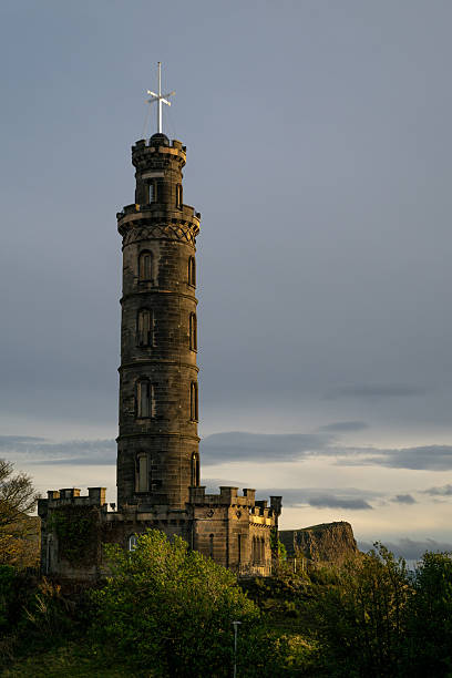 The Nelson Monument on Calton Hill in Edinburgh is pictured at golden hour, with soft sunlight illuminating the stone tower and surrounding greenery.