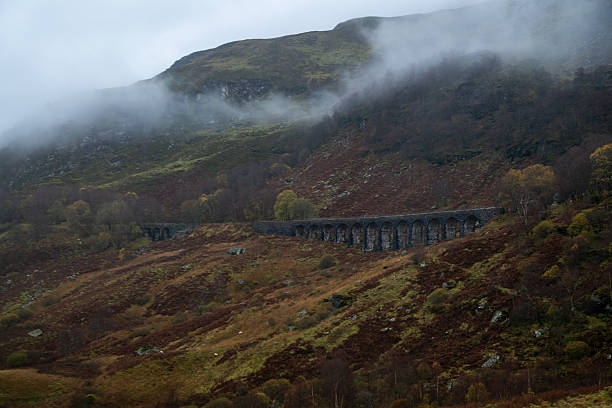 A long stone railway viaduct stretches across a misty hillside in the Scottish Highlands, surrounded by autumn colours and low clouds.