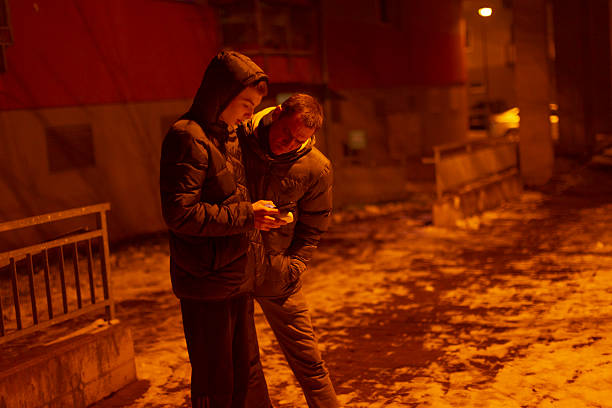Two white men huddled over phone display beside construction barrier and snowy pavement, close body language, shared focus on solutions and onscreen information, nighttime amber lighting and intimate
