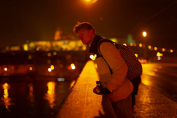 Young man photography, Male photographer standing on bridge overlooking water and illumination, Young man dressed in warm jacket capturing scenic reflections and city brightness at night