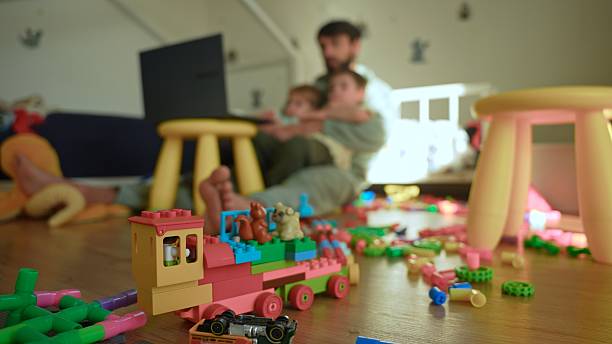 Messy Floor with Toys in Focus while a Father and Two Sons are Sitting in the Background Using a Laptop Together,Representing Modern Parenting,Work-Life Balance and Childhood
