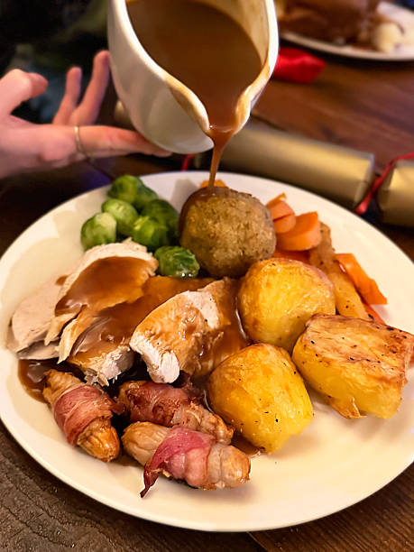 Image of unrecognisable woman pouring jug of gravy over plate containing Christmas dinner, slices of turkey meat, pigs in blankets, roasted potatoes, carrots, stuffing ball, Brussel sprouts, gold Christmas cracker, elevated view, focus on foreground