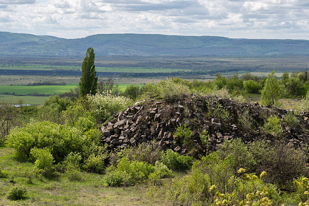 A mound of weathered basalt rocks is surrounded by lush spring shrubs overlooking a wide valley. In the distance, layered hills and fields stretch into the horizon under a softly clouded sky, creating a peaceful natural scene.