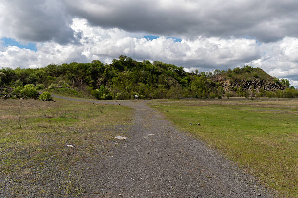 A gravel path leads toward a basalt hillside covered in lush spring vegetation under a dramatic cloudy sky. The open grassy field in the foreground contrasts with the rugged rock formations in the distance, creating a calm yet striking natural scene.