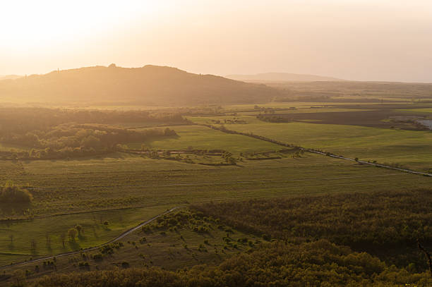 A warm golden sunset illuminates a vast rural valley with fields, meadows, and patches of forest. Soft light creates gentle contrasts across the landscape, highlighting the distant hills and tranquil natural scenery.