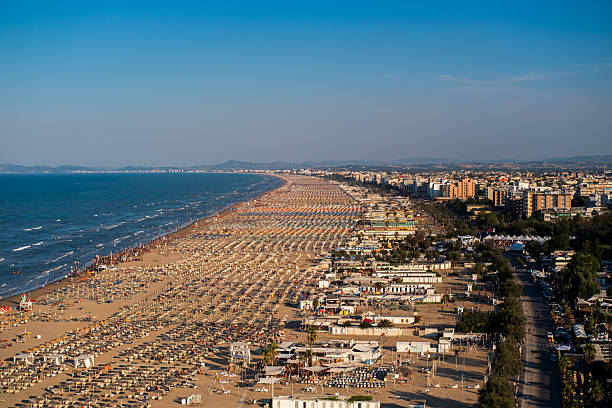 Aerial view of a packed sandy beach lined with umbrellas and sunbathers, stretching along a vibrant coastal city under clear skies. Rimini, Italy.