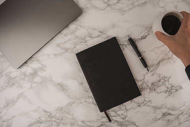 istock A hand holds a coffee cup while a black notebook and pen lie next to a laptop on a marble table. This scene captures a calm morning workspace atmosphere perfect for productivity 2250354685