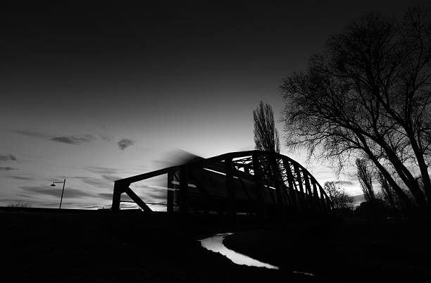 Old rusty truss bridge with moving freight train over the Orljava river. Moving train late in the evening. Rays of light. Evening arrival of the train. Black & White