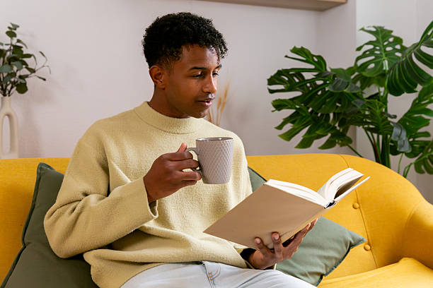 istock Young man relaxing on couch, reading book and drinking coffee 2250362927