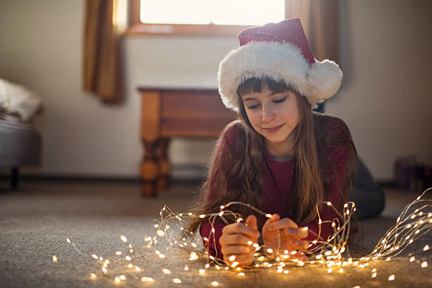 Cute little girl lies on the floor, smiling as she untangles a bundle of glowing string lights, preparing it for Christmas tree. The warm glow of the lights reflects on her face, adding to the cozy atmosphere of the room. Shot with Nikon D800