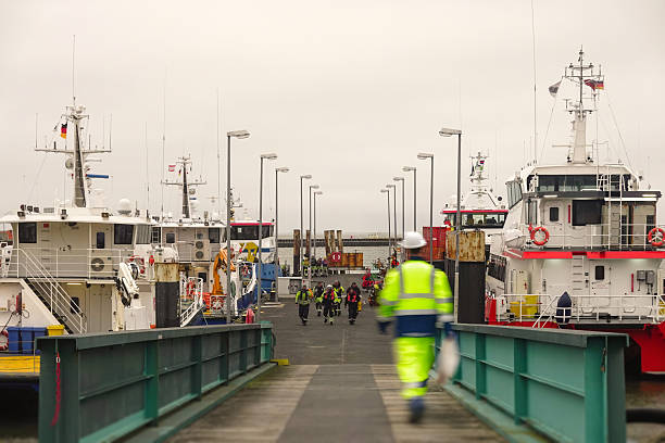 A wide, symmetrical shot capturing a team of marine personnel in high-visibility safety gear walking off a gangway and onto a port pier. They are flanked by multiple large, modern offshore service vessels docked in a harbor under an overcast sky. This image emphasizes the onshore logistics, preparation, and crew transfer procedures necessary to support offshore industrial operations.