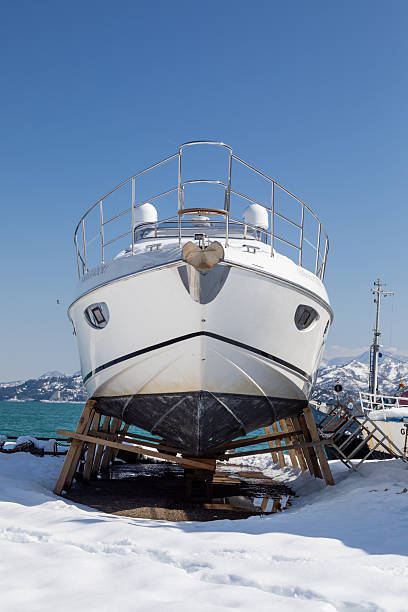 A blue-hulled boat rests on supports in a wet marina yard, with other vessels and a green pole visible under an overcast sky.