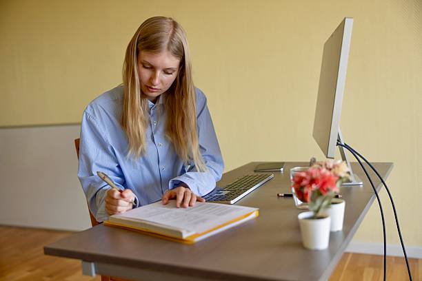 istock Young Gen Z woman sitting at a desk writing notes on paper while working or studying in a bright home office with a computer monitor 2250374620