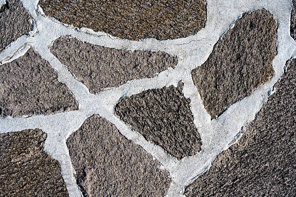 A detailed close-up of a rustic stone wall featuring irregular natural stones set in light-colored mortar. The textured surface shows rough, weathered rock with organic shapes and earthy tones.