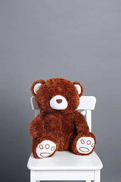 istock A brown teddy bear sits on a white chair against a gray wall. 2250412605