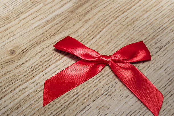 Shiny red satin ribbon is tied into a delicate bow knot resting on a light brown wood grain surface. Symbolizing a special gift, love or important memory. Great for gift concepts.