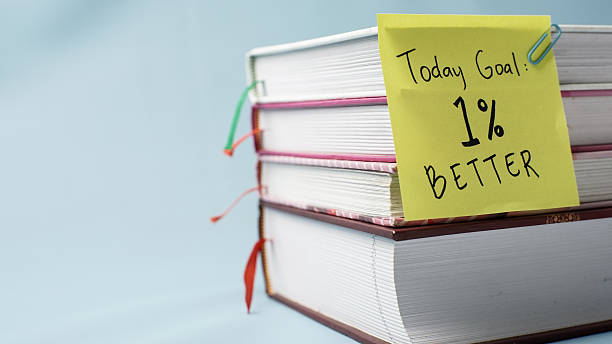A stack of books with a yellow sticky note reading “1% Better” clipped to the cover, set against a clean blue background for a motivational study or productivity theme.