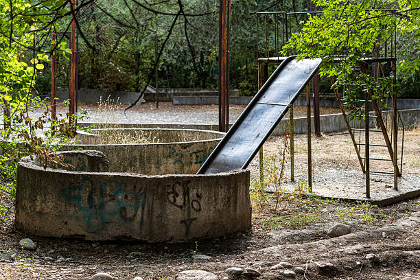 Bishkek, Kyrgyzstan - September 19, 2025: An old, abandoned, and neglected children's playground featuring a rusty metal slide and dilapidated concrete structures, overgrown with vegetation. The scene evokes a sense of decay, nostalgia, and forgotten memories.