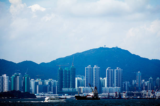 A coastal city with boats, cranes, high-rises, and forested mountains, showing urban growth and maritime activity.