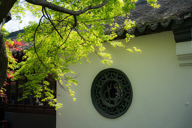 A traditional Chinese building features a circular window with intricate design, surrounded by green leaves and branches.