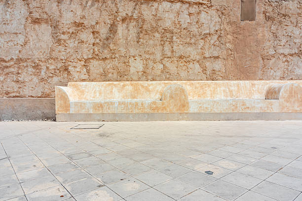 A weathered stone bench lines a sunlit courtyard against a rough, earthy wall. The scene conveys calm, solitude, outdoor seating, and rustic urban texture with warm beige tones.