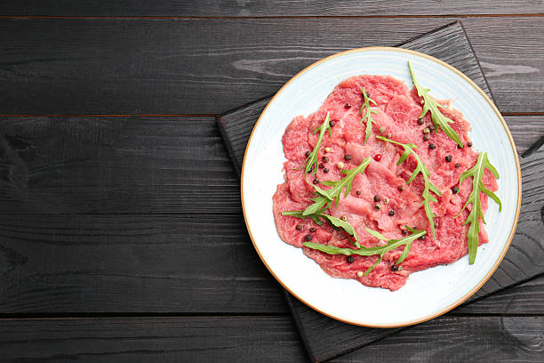 Fresh beef carpaccio with arugula and peppercorns on black wooden table, top view. Space for text