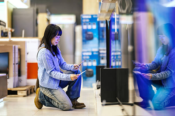 Young adult woman kneeling in an electronics store, comparing product specifications and prices on her smartphone and a paper flyer while shopping for new technology