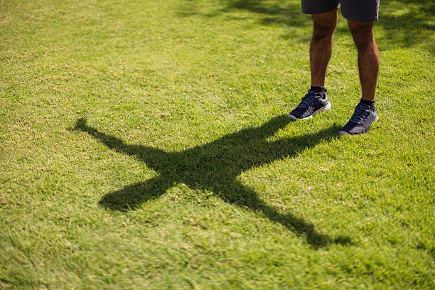 A man stands on lush green grass in bright sunlight, casting a distinct long shadow. The focus is on his legs and blue athletic shoes, suggesting outdoor activity, fitness, or a casual exercise scene in a park-like setting.