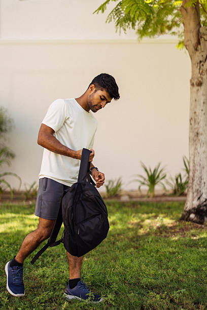 A fit young man in a light t-shirt and shorts stands on the grass beside a tree, adjusting a black backpack strap. The outdoor scene is bright and calm, with soft light and a neutral background, evoking travel, fitness, and everyday commuting moments.