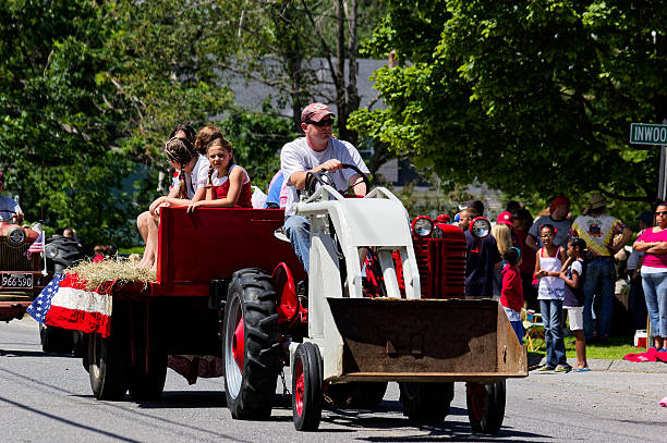 RUTLAND, MA - July 04, 2009: Family enjoys Independence Day parade riding vintage tractor with large rear wheels. Three children wave American flags from red hay-filled wagon as spectators cheer from