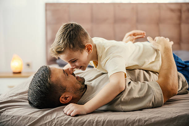 Father and elementary-age son laugh and bond while playfully tickling each other on a cozy bed at home, sharing relaxed, loving weekend family time and joyful connection