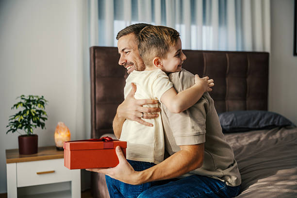 Father and son embrace in a cozy bedroom as the child receives a wrapped gift, sharing a tender, joyful moment of celebration, love and gratitude during the day