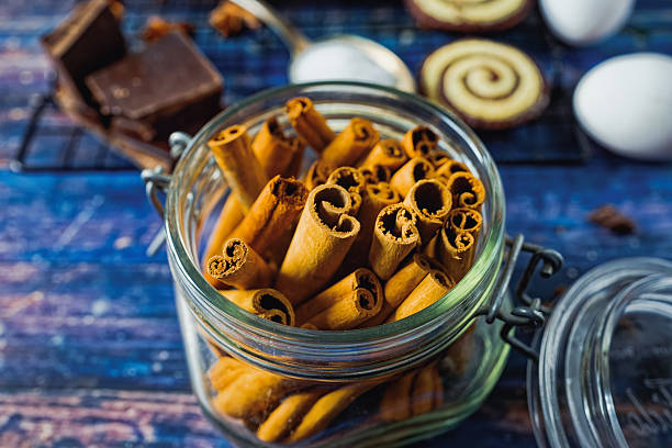 Close-up of a glass jar filled with cinnamon sticks on a rustic blue wooden background, surrounded by baking ingredients such as chocolate and cookies in soft focus. The warm tones and natural textures evoke a cozy, homemade baking atmosphere. Ideal for themes of slow living, sustainability, and seasonal cooking. Copy space appears around the edges of the frame.