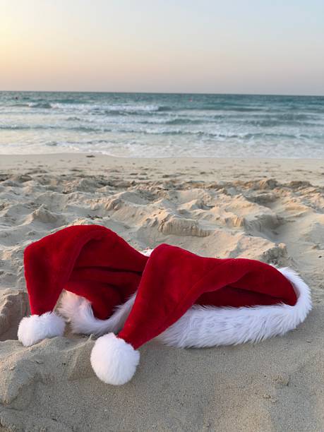 istock Christmas Santa hats on sandy beach by ocean waves 2250496381