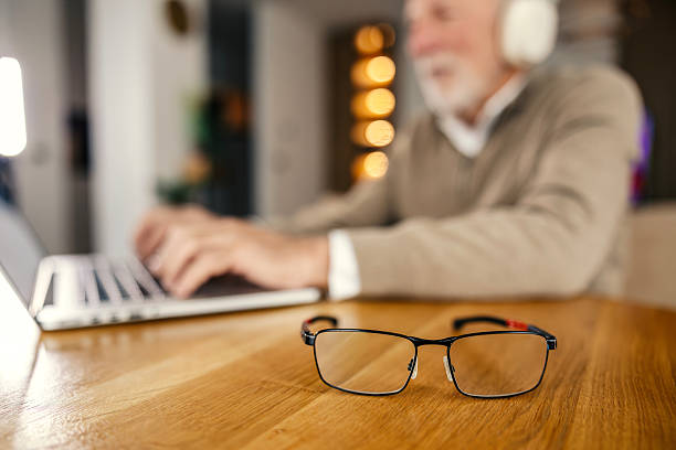 Reading glasses are resting on a wooden table with a blurred senior man wearing headphones and typing on a laptop in the background, focusing on his remote work