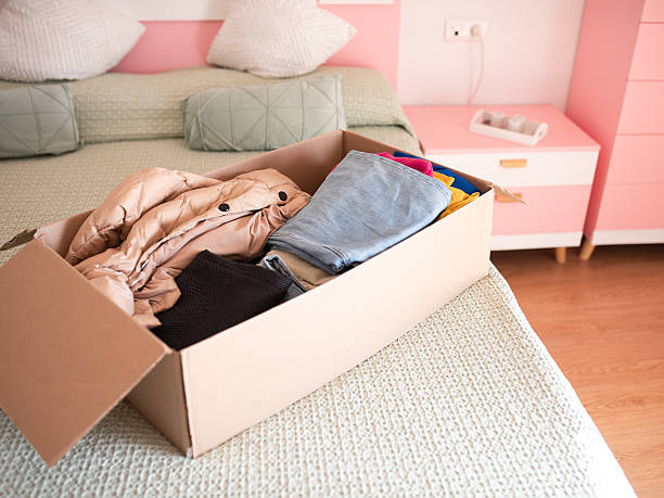 istock Cardboard box filled with assorted clothing items, including jackets and pants, placed on a neatly made bed in a cozy bedroom setting with pastel colors 2250498999