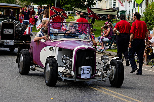 Rutland, Ma - July 04, 2009: Vintage hot rod cruises down suburban street in patriotic parade, passengers waving both Confederate and American flags from open-top car. Crowd in red shirts lines street