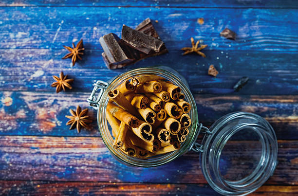 Overhead view of a glass jar filled with cinnamon sticks placed on a rustic blue wooden table, surrounded by star anise and dark chocolate pieces. The warm spices and textured background create a cozy, seasonal baking atmosphere. Ideal for themes of homemade cooking, natural ingredients, and winter baking traditions. Copy space is present around the upper and side areas of the frame.