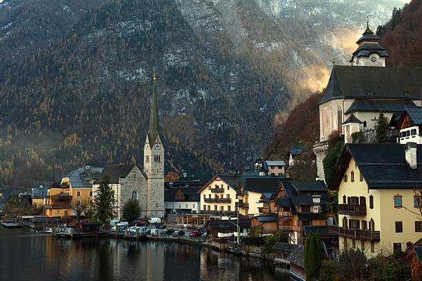 A serene lakeside village rests beneath a forested mountain, featuring alpine homes, a clock-towered church, and tranquil reflections—capturing the charm and peaceful beauty of a European mountain.